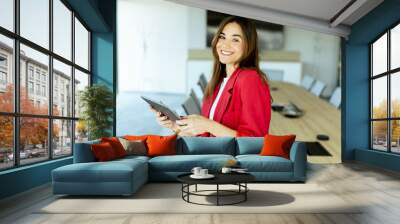 Smiling professional woman in red blazer holding tablet in modern conference room during daytime Wall mural