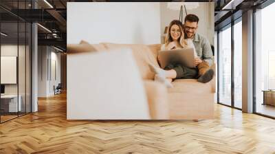 Young couple using laptop together while sitting on sofa at home Wall mural