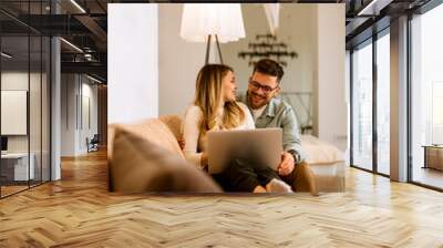 Young couple using laptop together while sitting on sofa at home Wall mural