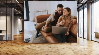 Young couple using laptop together while sitting on sofa at home Wall mural