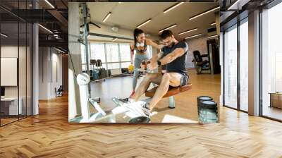 Young man using seated row machine in the gym with support of female coach Wall mural