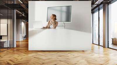 Young woman working on laptop in bright office with big screen behind her Wall mural