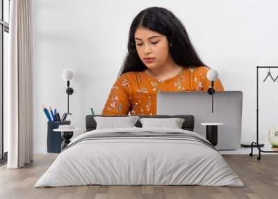 Pretty young Indian woman sitting against white backdrop and using her laptop. Cute teenager doing her online schoolwork from home. Wall mural