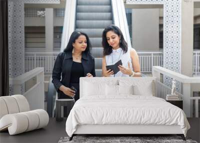 Two smiling  Indian women looking into a phone as they come down an escalator in an urban corporate setting, Wall mural