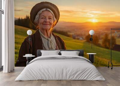 Smiling senior farmer woman standing on a meadow in the countryside at a sunset and looking at the camera. Wearing old village clothes. Sunset over the green pasture in the background Wall mural