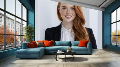 Young Caucasian woman with red hair and freckles smiling in a black suit jacket and white shirt leaning against a white wall in a bright office setting for a professional business portrait Wall mural