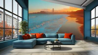 A panoramic view of a sandy beach with waves crashing against the shore at sunset, with a city skyline in the distance. Wall mural