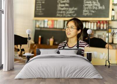 Asian girl with glasses waitress wearing apron standing in coffee shop. Wall mural