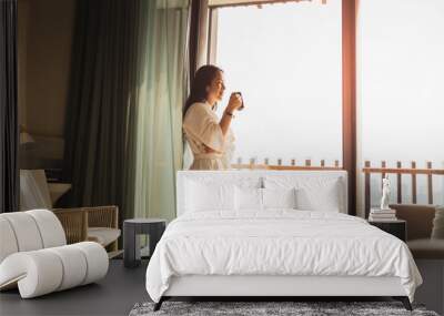 Happy businesswoman in dressing gown stands near the window holding cup of coffee. Wall mural