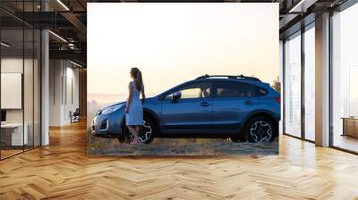 Happy young woman driver in blue dress enjoying warm summer evening standing beside her car. Travelling and vacation concept Wall mural