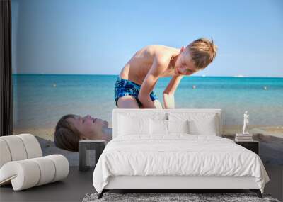 Two kids play on beach, laughing boy covers smiling girl with white sand, bright blue sky and sea water background Wall mural