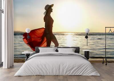 Young woman wearing long red dress and straw hat standing on sand beach at sea shore enjoying view of rising sun in early summer morning. Wall mural