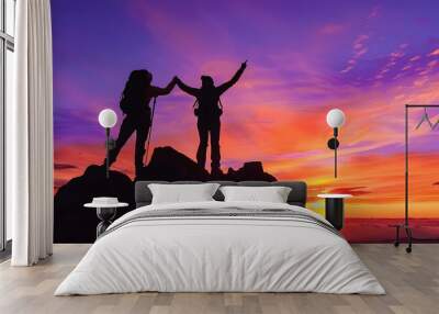 Silhouettes of two hikers against an orange and purple sky at sunset, celebrating their achievement atop a lofty mountain Wall mural