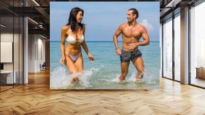 Happy couple laughing together holding hands running having fun splashing water in the ocean waves. Young beautiful fit slim body people enjoying their happy lifestyle in paradise destination beach. Wall mural