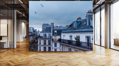 A captivating scene captures the view from a fifth-floor balcony overlooking Parisian rooftops before nightfall. Chimneys, rooftops, and numerous windows adorn the landscape, illuminated windows Wall mural