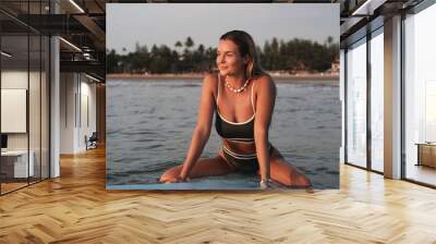 Young woman in a bikini sits on her surfboard in shallow ocean water Wall mural