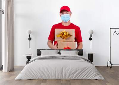 Safe contactless remote delivery of holiday gifts during the coronavirus pandemic. A courier in a red uniform and protective medical mask holds two beautiful gift boxes with bows Wall mural