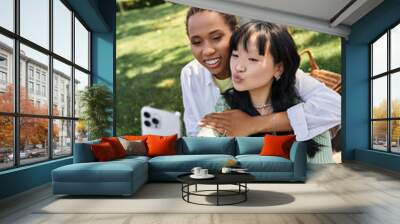 A lesbian couple enjoys a picnic in the park, taking a selfie with a smartphone. Wall mural