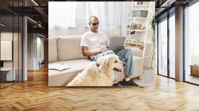 A man with myasthenia gravis syndrome works on a laptop while a loyal Labrador dog keeps him company on the couch. Wall mural