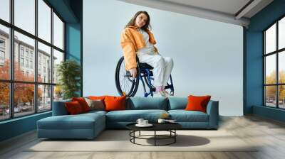 A young brunette woman in a wheelchair and stylish casual attire poses against a grey backdrop. Wall mural