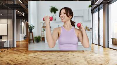 A young woman in a purple tank top smiles while lifting pink weights in a modern apartment with white cabinets. Wall mural