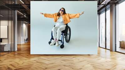 A young woman in a wheelchair wears an orange jacket and white pants, with arms outstretched in freedom and happiness Wall mural