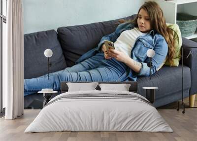 A young woman relaxes on a couch in her modern apartment, scrolling through her phone. Wall mural