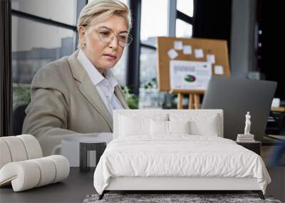 An adult woman dressed elegantly engages with her laptop at a contemporary office desk. Wall mural