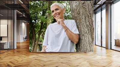 Mature woman smiles while leaning against a tree, embracing a sunny day outdoors. Wall mural