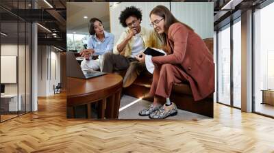 Three colleagues, including a woman with a prosthetic leg, discuss a project on a tablet while sitting on a couch in a modern office lobby. Wall mural