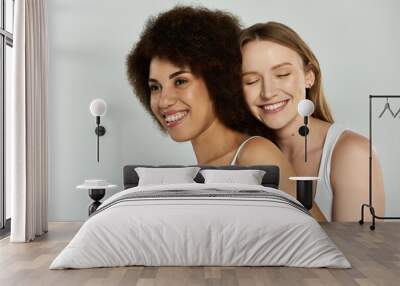 Two women in white tank tops and jeans smile at each other against a grey background, expressing their love and joy as a couple Wall mural