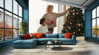 Two women share laughter and love while decorating a cheerful Christmas tree in their cozy home. Wall mural