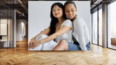 Two young women, one African American and one Asian, embrace on a grey backdrop. Wall mural