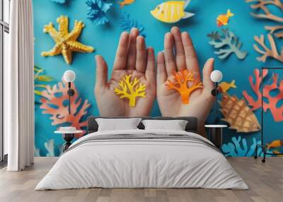 Two hands holding paper cut vibrant coral pieces, one yellow and one orange, with a colorful blue background filled with various corals, showcasing the beauty and diversity of marine life. Wall mural