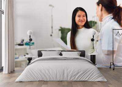 A smiling Asian female patient talking with a female doctor, sitting in the medical office. Wall mural