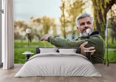 A sporty senior bearded man stretches his arms in nature in the morning. Wall mural