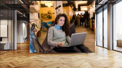 Beautiful young woman working on laptop and smiling while sitting indoors. Wall mural