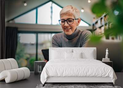 Close-up portrait of a happy senior woman looking at smart phone while sitting at desk with laptop. Wall mural