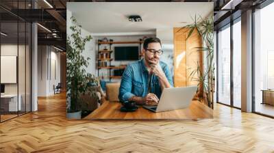 Portrait of a casual businessman thinking while looking at laptop screen, keeping hand on chin. Wall mural