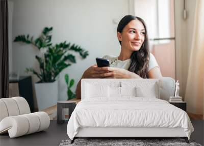 Portrait of a casual young woman using smart phone at home. Looking away. Wall mural