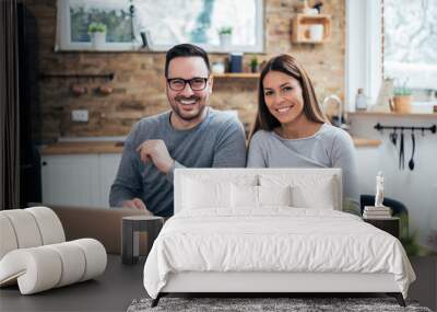 Portrait of a cheerful young couple at the kitchen table with laptop and breakfast, looking at camera. Wall mural