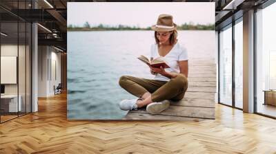 Portrait of a girl reading a book while sitting on a small wooden wharf. Wall mural