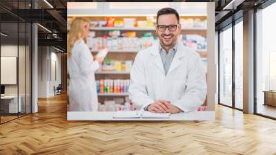 Portrait of a handsome pharmacist at the counter of a drugstore, female colleague working in the background. Wall mural