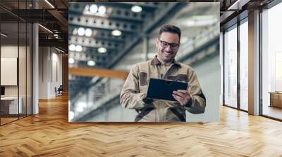 Portrait of a smiling factory engineer, low angle image. Handsome man looking at tablet while working at industrial building. Wall mural