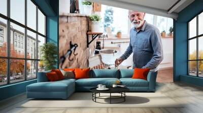 Portrait of a smiling senior man cooking food at home, looking at camera. Wall mural