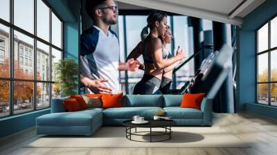 Side view of fit young man and women running side by side on treadmills at the gym. Wall mural