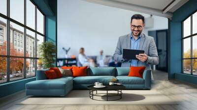 Smiling businessman using a tablet while standing, dressed elegantly. Wall mural