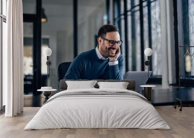 Smiling man with glasses on, using a laptop, at his office. Wall mural