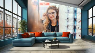 smiling young woman trying on glasses on mirror in optician. Wall mural