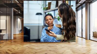 Two females enjoying their coffee break, looking at a mobile phone, watching something online. Wall mural